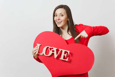 Young pretty smiling woman in red casual clothes pointing on big red heart, wooden word love on white background. Copy space for advertisement. St. Valentines Day or International Women's Day conceptの写真素材