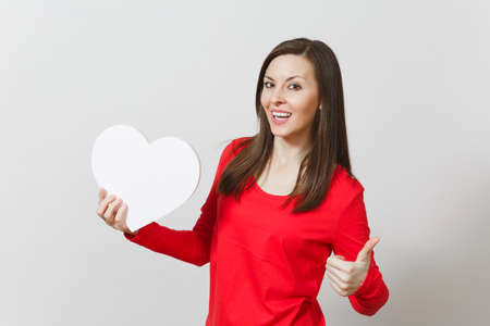 Pretty woman in red clothes showing thumb up, holding big white heart in hands isolated on white background. Copy space for advertisement. St. Valentine's Day or International Women's Day conceptの写真素材