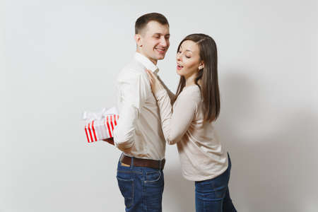 Cute fun couple in love. Woman looking behind man who hiding behind him red present box with gift isolated on white background. St. Valentine's Day, International Women's Day birthday holiday conceptの写真素材