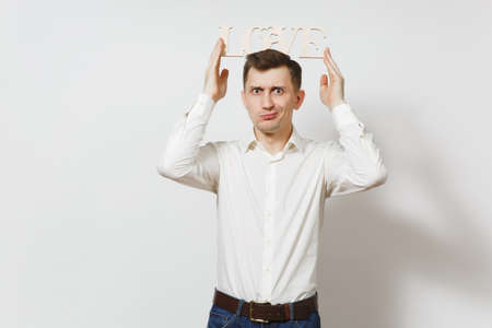 Young confused man in shirt holding above head wooden word Love isolated on white background. Copy space for advertisement. St. Valentine's Day, International Women's Day, birthday, holiday conceptの写真素材