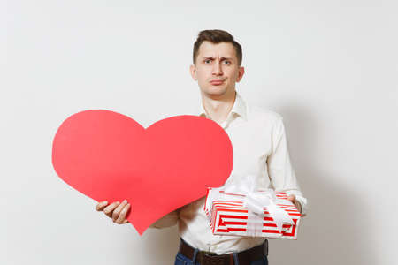 Dissatisfied man in shirt holding big red heart, present box with gift isolated on white background. Copy space for advertisement. St. Valentines Day, International Women Day birthday holiday conceptの写真素材