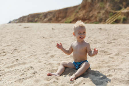 Little smiling cute child baby boy sitting at sea on sand summer beach, mountain background, looking on water. Little kid son playing outdoors. Family day 15 of may, love, parents, children conceptの写真素材