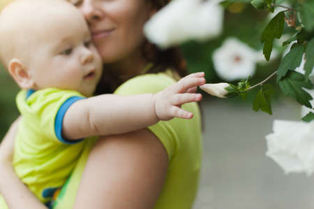 Young tender woman standing at green bush hugging little cute child baby boy stretching hand to white flowers. Mother, little kid son. Parenthood, family day 15 of may, love, parents children conceptの写真素材