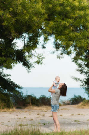 Young fun smiling woman standing at sea background hugging little cute child baby boy on nature, green trees. Mother, little kid son. Parenthood, family day 15 of may, love, parents, children conceptの写真素材
