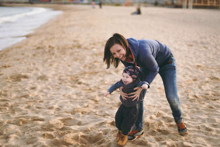 Young woman playing at sea sand beach background with little cute child baby boy, good mood. Mother, little kid son on sunny day. Parenthood, family holiday 15 of may, love, parents, children conceptの写真素材