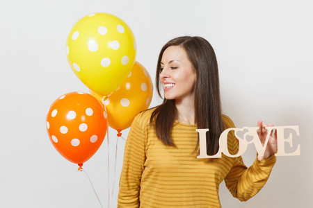 Beautiful romantic young smiling woman holding wooden word love, yellow orange air balloons on white background. Copy space for advertisement. St. Valentine's Day or International Women's Day conceptの写真素材
