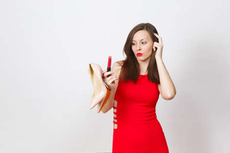 Beautiful glamour caucasian fashionable young brown-hair woman in red dress holding beige shoes with red sole, lipstick of her shopping isolated on white background. Copy space for advertisementの写真素材