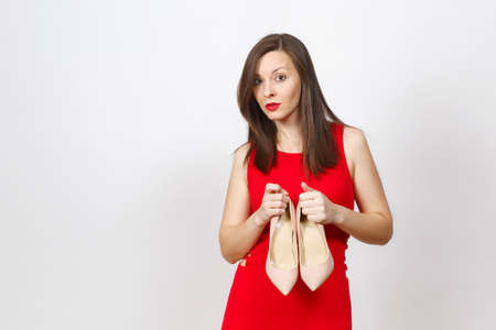 Smiling glamour caucasian fashionable young smiling brown-hair woman in red dress holding pair of beige shoes with red sole of her shopping isolated on white background. Copy space for advertisementの写真素材