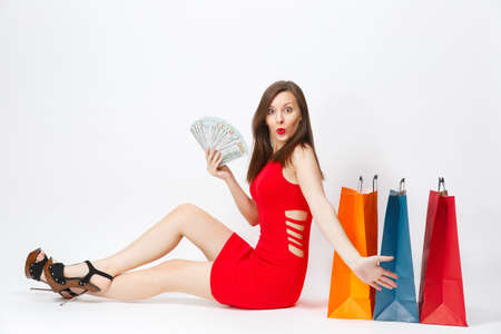 Beautiful glamour fashionable young woman in red dress sitting with cash dollars, multi colored packets with purchases after shopping isolated on white background. Copy space for advertisementの写真素材