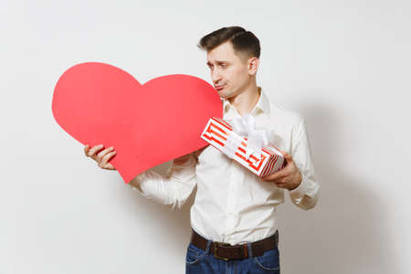 Dissatisfied man in shirt holding big red heart, present box with gift isolated on white background. Copy space for advertisement. St. Valentines Day, International Women Day birthday holiday conceptの写真素材