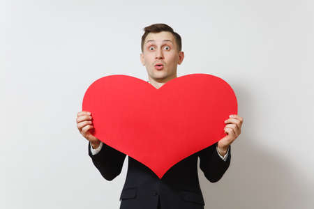 Young stunned shocked upset sad man in suit holding big red heart isolated on white background. Copy space for advertisement. St. Valentine's Day, International Women's Day, birthday, holiday conceptの写真素材