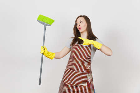 Young smiling housewife in striped apron, yellow gloves isolated on white background. Fun housekeeper woman cleaning maid holding and sweeping with broom Copy space for advertisement Advertising areaの写真素材