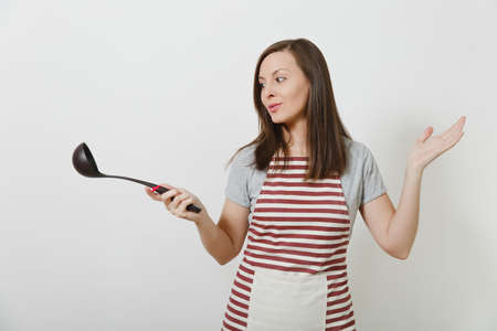 Young attractive smiling caucasian housewife in striped apron isolated on white background. Beautiful housekeeper woman holding black kitchen soup ladle. Copy space for advertisementの写真素材