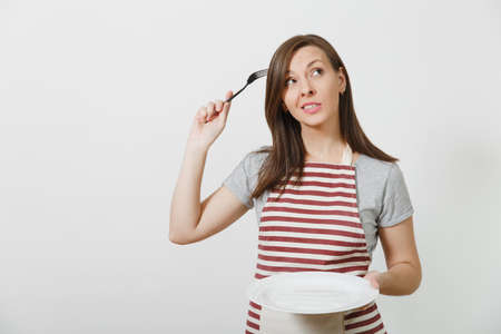 Young fun crazy brunette housewife in striped apron gray t-shirt isolated on white background Housekeeper woman holding white empty plate fork in hair like comb hairbrush. Copy space advertisementの写真素材