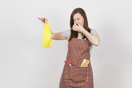Young sad upset tired shocked housewife in striped apron with cleaning rag in pocket isolated on white background. Housekeeper woman holds smell yellow gloves covers nose with hand. For advertisementの写真素材