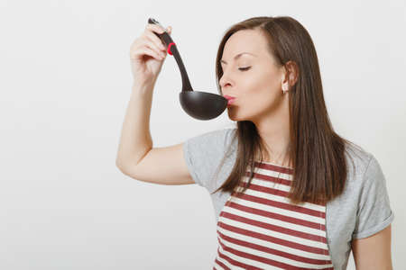 Young attractive smiling caucasian housewife in striped apron isolated on white background. Beautiful housekeeper woman tries sniffs enjoys aroma soup from black kitchen soup ladle. Delicious tasteの写真素材