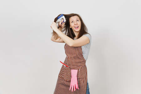 Young fun crazy dizzy loony wild housewife in striped apron with squeegee, pink gloves in pocket isolated on white background. Mad woman comb out tousled brown hair by brush for cleaning. Copy spaceの写真素材