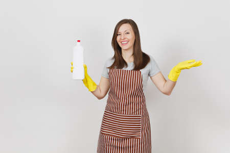 Young housewife in yellow gloves, striped apron, cleaning rag in pocket isolated on white background. Woman pours cleaner liquid for washing dishes from bottle on sponge. Copy space for advertisementの写真素材