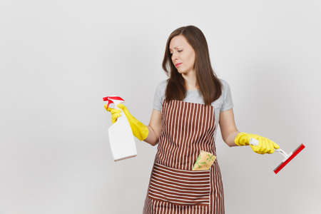 Young sad upset bewildered housewife in yellow gloves striped apron cleaning rag squeegee in pocket isolated on white background. Woman spread hands, hold spray bottle with cleaner liquid. Copy spaceの写真素材