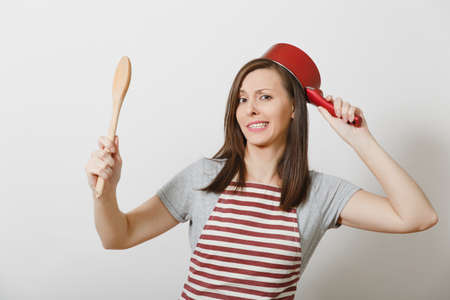 Young smiling crazy housewife in striped apron, gray t-shirt isolated on white background. Beautiful fun housekeeper woman holding red empty stewpan at head wooden spoon. Copy space for advertisementの写真素材