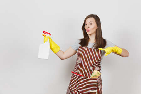 Young smiling housewife in yellow gloves, striped apron, cleaning rag, squeegee in pocket isolated on white background. Housekeeper woman holding spray bottle with cleaner liquid. Bottle copy spaceの写真素材