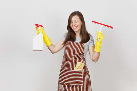 Young smiling housewife in yellow gloves, striped apron, cleaning rag in pocket isolated on white background. Housekeeper woman holding squeegee, spray bottle with cleaner liquid. Bottle copy spaceの写真素材