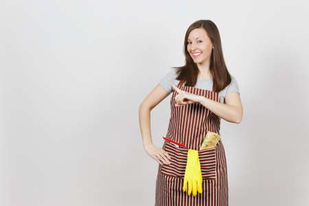 Young smiling housewife in striped apron with cleaning rag, squeegee, yellow gloves in pocket isolated on white background. Beautiful housekeeper woman pointing index finger aside looking cameraの写真素材