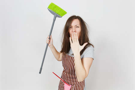 Young fun crazy dizzy loony wild screaming housewife tousled hair in striped apron isolated on white background. Mad witch woman with broom, shut up, cover mouth by hand. Copy space for advertisementの写真素材