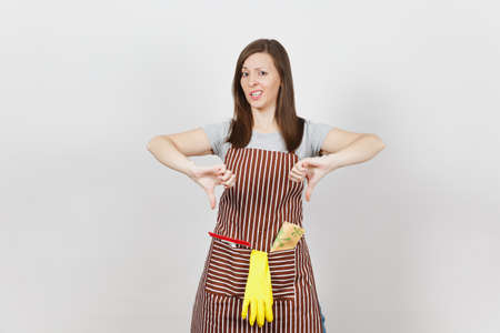 Young sad upset tired crying housewife in striped apron with cleaning rag, squeegee, yellow gloves isolated on white background. Beautiful housekeeper woman showing thumbs down. For advertisementの写真素材