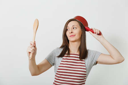 Young smiling crazy housewife in striped apron, gray t-shirt isolated on white background. Beautiful fun housekeeper woman holding red empty stewpan at head wooden spoon. Copy space for advertisementの写真素材