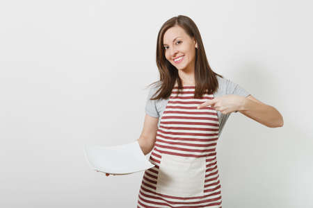 Young attractive smiling brunette caucasian housewife in striped apron, gray t-shirt isolated on white background. Beautiful housekeeper woman holding white empty plate. Copy space for advertisementの写真素材