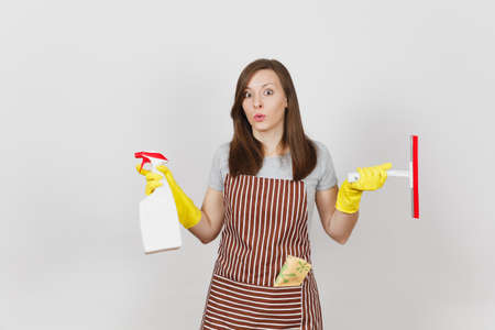 Young sad upset bewildered housewife in yellow gloves striped apron cleaning rag squeegee in pocket isolated on white background. Woman spread hands, hold spray bottle with cleaner liquid. Copy spaceの写真素材