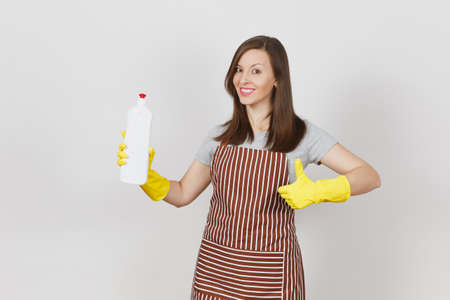 Young smiling housewife in yellow gloves, striped apron isolated on white background. Woman showing thumb up, holding bottle with cleaner liquid for washing and cleaning. Copy space for advertisementの写真素材