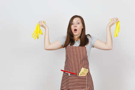 Young sad upset tired shocked housewife in striped apron with cleaning rag in pocket isolated on white background. Pretty housekeeper woman holding yellow gloves in spreading hands. For advertisementの写真素材