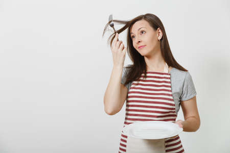 Young fun crazy brunette housewife in striped apron gray t-shirt isolated on white background Housekeeper woman holding white empty plate fork in hair like comb hairbrush. Copy space advertisementの写真素材
