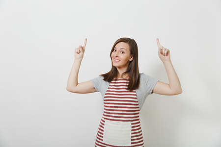 Young attractive smiling brunette caucasian housewife in striped apron isolated on white background. Beautiful housekeeper woman pointing index fingers up looking camera. Copy space for advertisementの写真素材