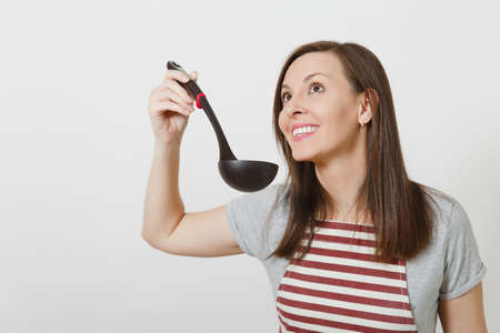 Young attractive smiling caucasian housewife in striped apron isolated on white background. Beautiful housekeeper woman tries sniffs enjoys aroma soup from black kitchen soup ladle. Delicious tasteの写真素材