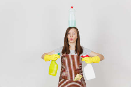 Fun housewife in yellow gloves, striped apron, cleaning rag in pocket on white background. Woman holding in hands, on head bottles with cleaner liquid for washing dishes. Copy space for advertisementの写真素材