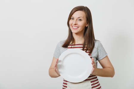 Young attractive smiling brunette caucasian housewife in striped apron, gray t-shirt isolated on white background. Beautiful housekeeper woman holding white empty plate. Copy space for advertisementの写真素材