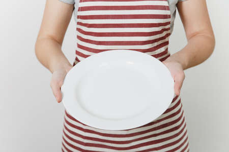 Young caucasian housewife in striped apron, gray t-shirt isolated on white background. Housekeeper woman holding in hands white empty round plate. Copy space for advertisementの写真素材
