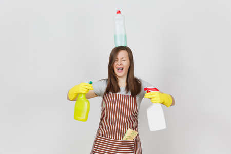 Fun housewife in yellow gloves, striped apron, cleaning rag in pocket on white background. Woman holding in hands, on head bottles with cleaner liquid for washing dishes. Copy space for advertisementの写真素材