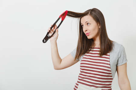 Young fun attractive smiling brunette caucasian housewife in striped apron isolated on white background. Beautiful housekeeper woman holding hair by kitchen tongs like styler curling hairの写真素材