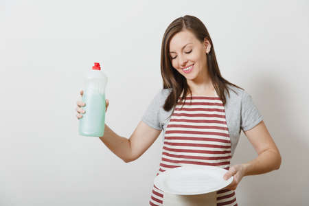 Young housewife in striped apron isolated on white background. Housekeeper woman holds bottle with cleaner liquid for washing dishes white empty round plate . Bottle with copy space for advertisementの写真素材