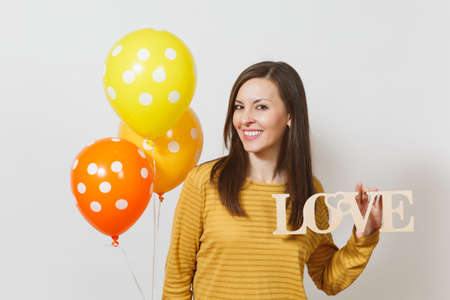 Beautiful romantic young smiling woman holding wooden word love, yellow orange air balloons on white background. Copy space for advertisement. St. Valentine's Day or International Women's Day conceptの写真素材