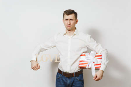 Young confused sad upset man in shirt holding armpit wooden word Love, present box with gift isolated on white background. St. Valentine's Day, International Women's Day, birthday, holiday conceptの素材 [FY31097829136]