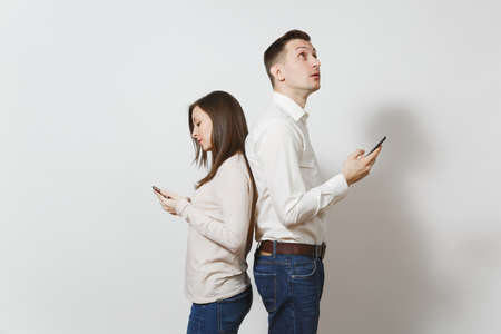 Young caucasian couple. Man and woman in love in casual light clothes, jeans standing back to back, using and texting on their mobile smart phones isolated on white background. Gadget today conceptの写真素材
