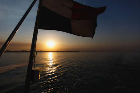 Beautiful sea landscape. Rectangular flag attached to the deck of the yacht on the background bright warm sunset in the sea near the coastline with the city's silhouettes.の写真素材