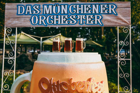 Treats for guests at the Oktoberfest. Three large glasses of fresh cold foam beer on a stand in the form of a cup under a breeching sign in the green park where the festival is held.の写真素材