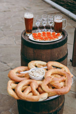 Treats for guests at the Oktoberfest. Salted soft baked fragrant pretzels and a spread for them in a plate with a knife, canapé with red caviar and glasses with drinks on a tray on a wooden barrels.の写真素材
