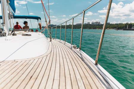 Sailing boat deck on a white yacht with teak wooden deck, metal railing, set of red ropes with people on board in the sea near the coastline in the background blue sky with clouds in sunny day.の写真素材